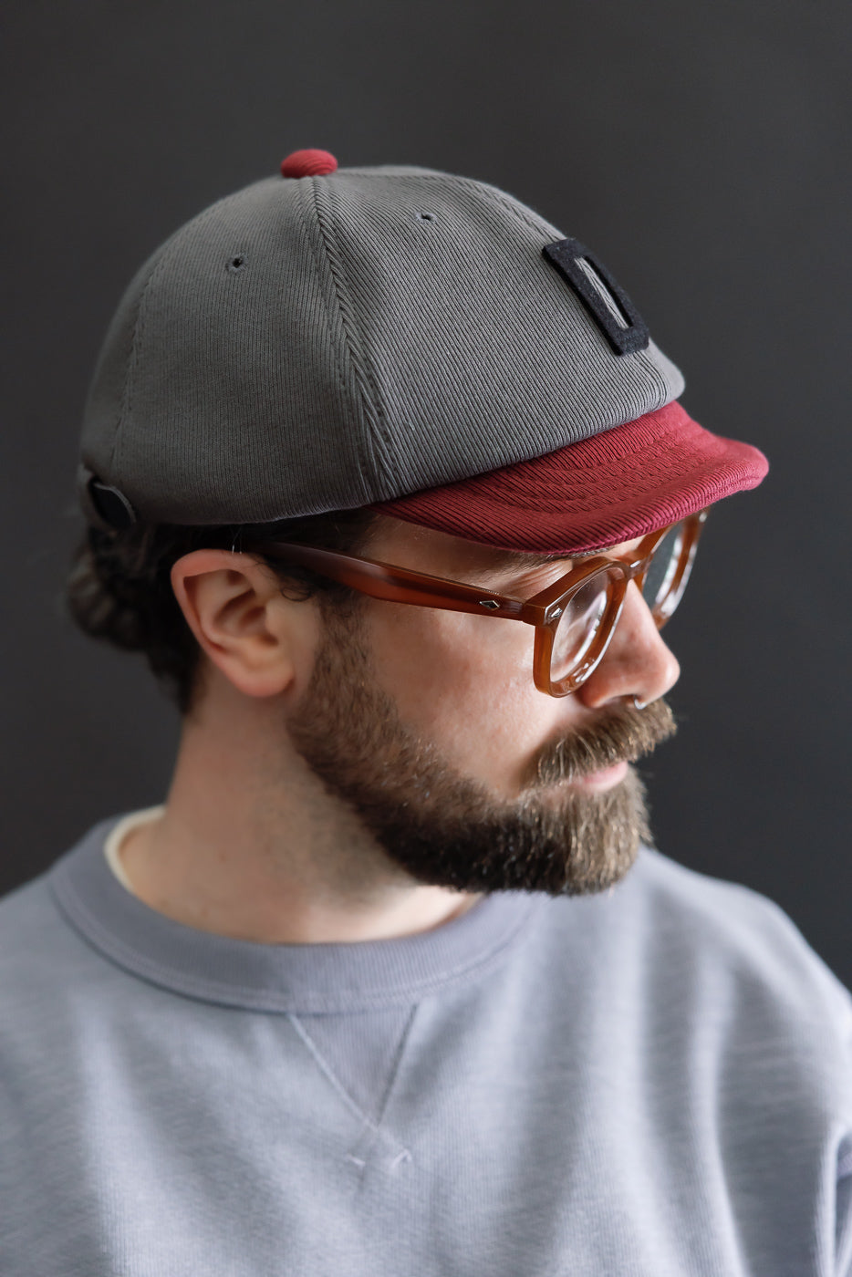 The JM6310 - GG Rib BB Cap D - 318 Gray x Oxblood features a two-tone design with a gray crown and burgundy brim, crafted from sustainable cotton ribbed fabric, showcased on a bearded model.