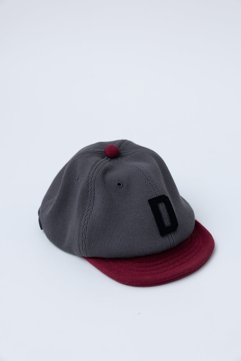 JM6310 - GG Rib BB Cap D - 318 Gray x Oxblood features a dark gray crown, burgundy visor, and a black D stitching, crafted sustainably from ribbed cotton and reclaimed materials.