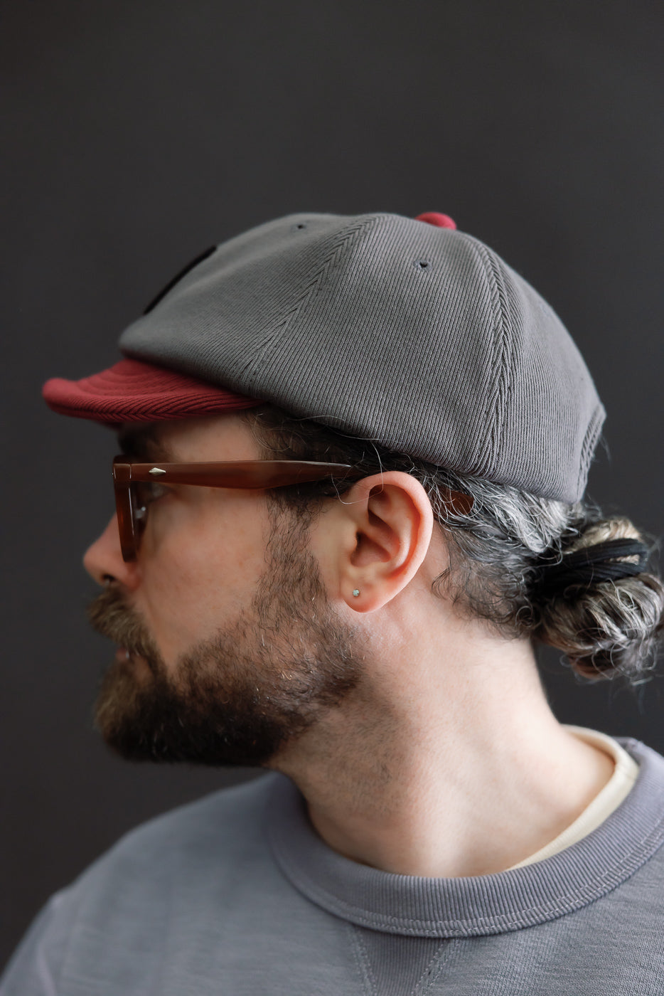 Person wearing JM6310 - GG Rib BB Cap D - 318 Gray x Oxblood, showcasing the cap's gray crown and reddish brim, with a low bun hairstyle and rectangular glasses, highlighting sustainable and comfortable design.