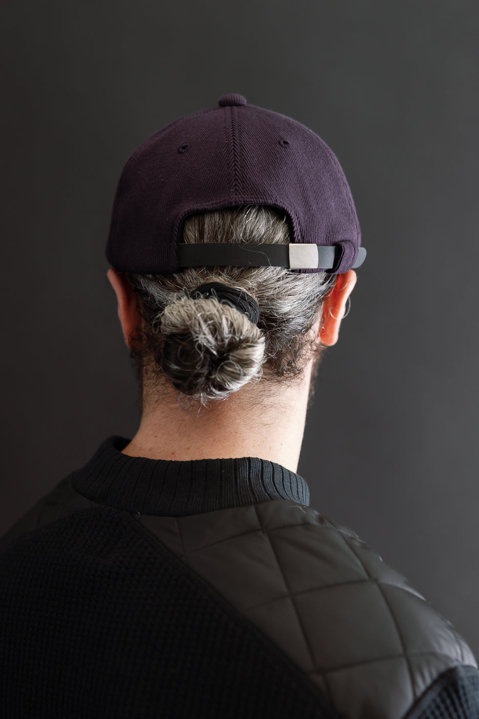 Back view close-up of a male model wearing a dark purple baseball cap and a black quilted sweater.