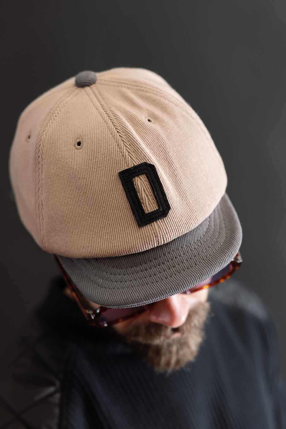Close-up front view of a tan corduroy baseball cap with a black letter D patch and grey brim.
