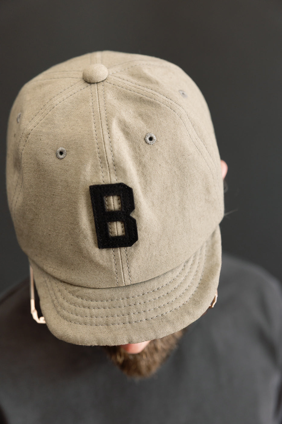 Close-up front view of beige cap with black 'B' patch, worn by male model, 5'9