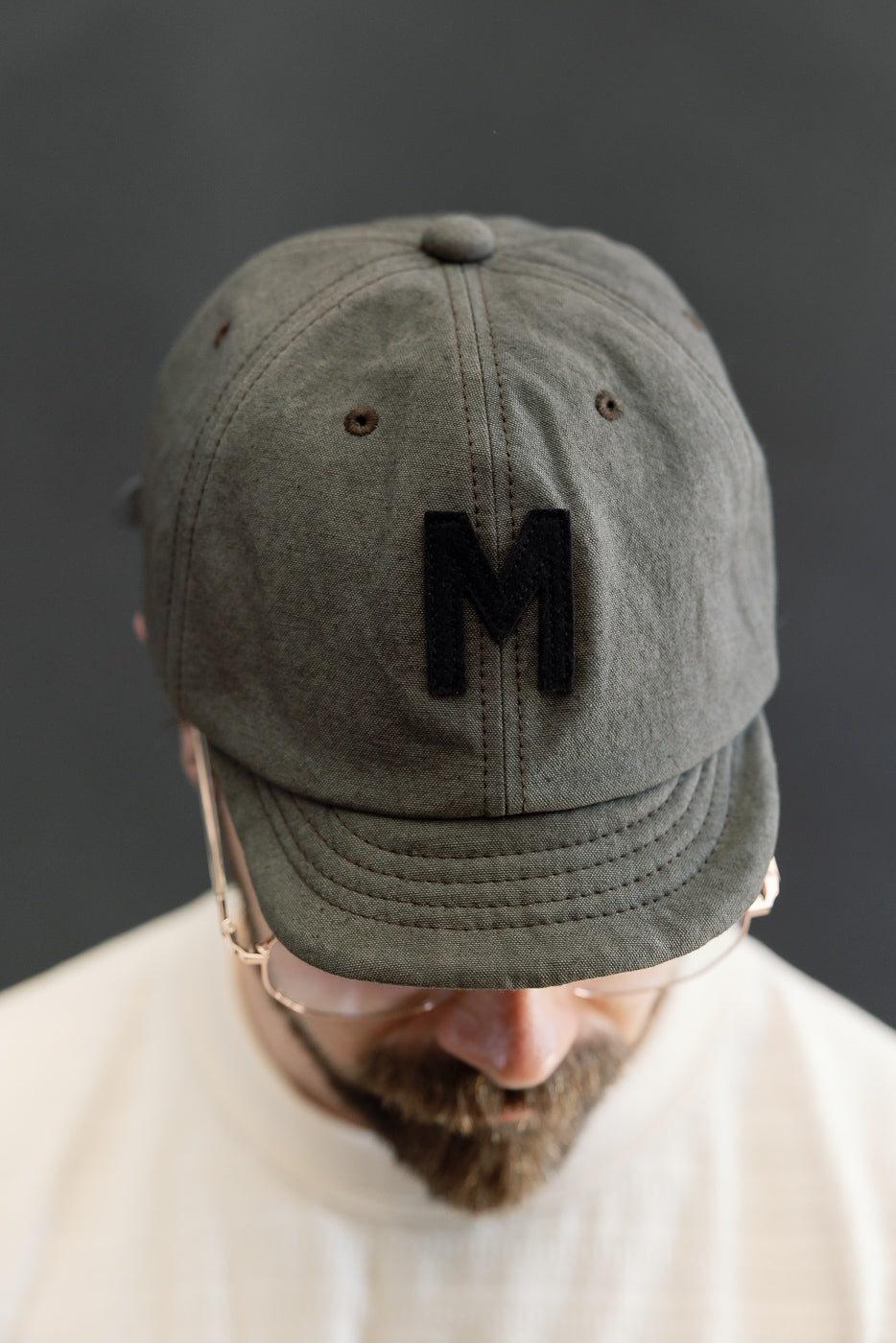 Close-up front view of dark moss gray canvas cap with black 'M' patch, worn by male model, 5'9