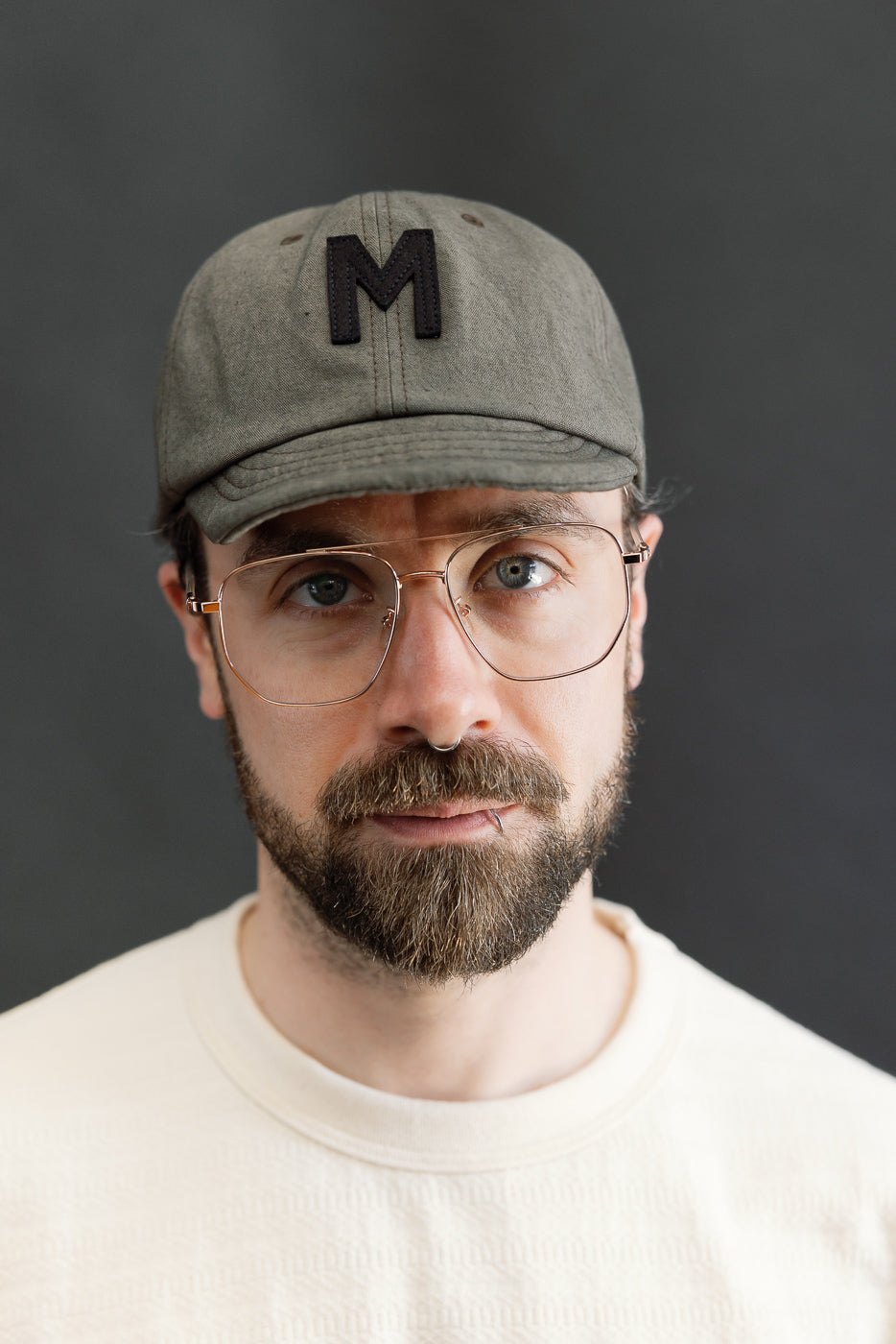 Close-up front view of dark moss gray canvas cap with black M patch on male model, 5'9