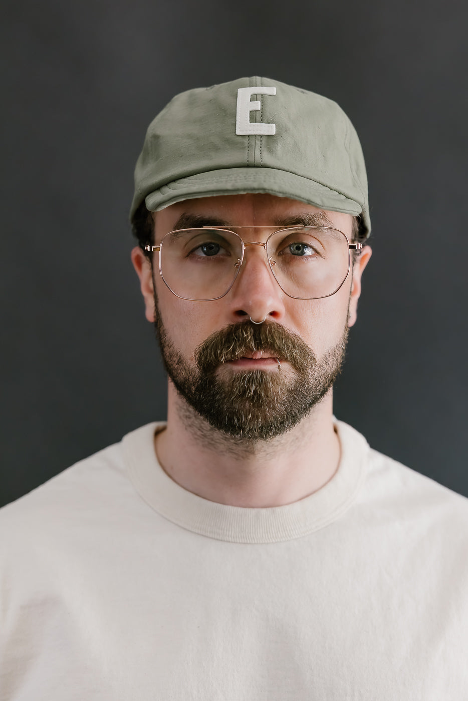 Man wearing a cap with a letter 'E' and glasses against a dark background