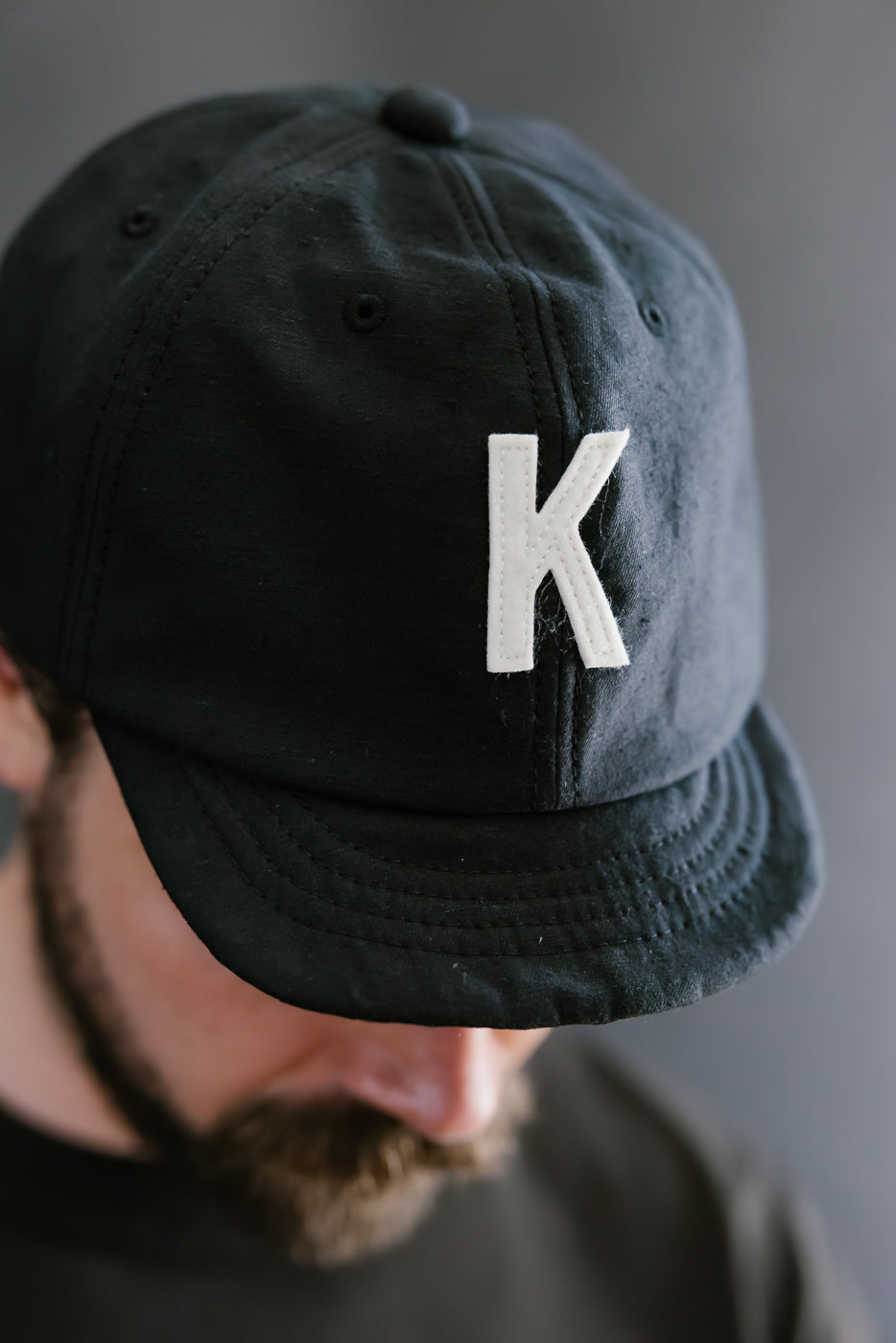 Black cap with a white 'K' logo worn by a person against a gray background