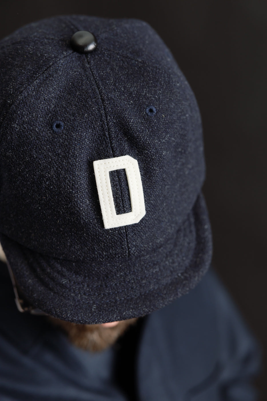 Close-up front view of dark navy wool baseball cap with white letter D, worn by male model, showing fabric texture and cap top details.