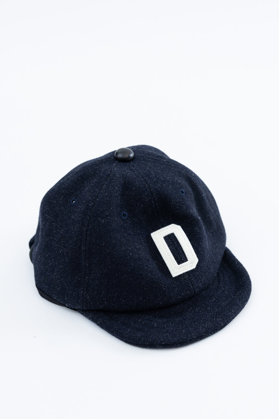 Close-up front view of dark navy melton wool baseball cap with white D patch and top button detail.