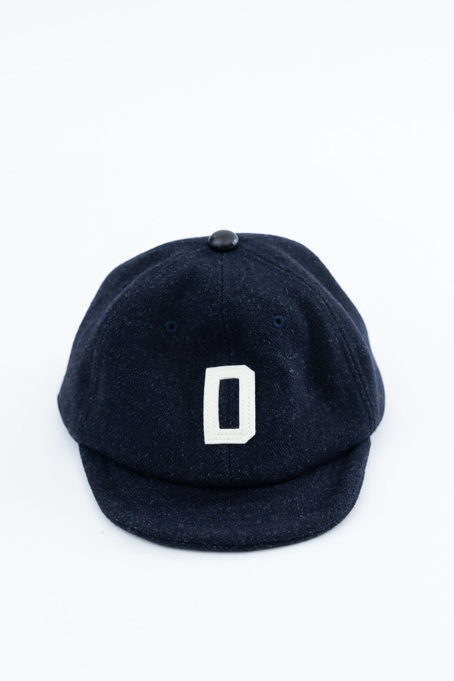 Close-up front view of dark navy melton wool baseball cap with white embroidered letter O and black top button.