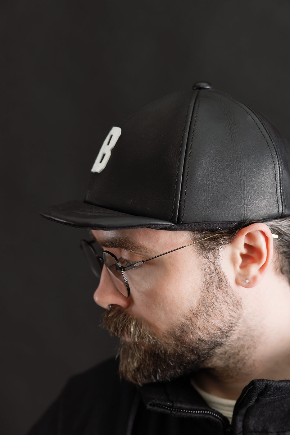 Close-up side view of black leather baseball cap with white B patch on male model, 5'9