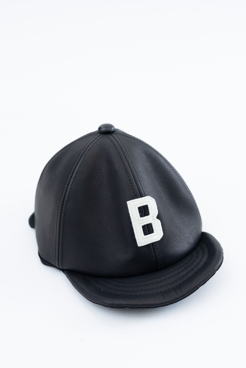 Front view close-up of black leather baseball cap with white embroidered B patch, curved brim, visible stitching.