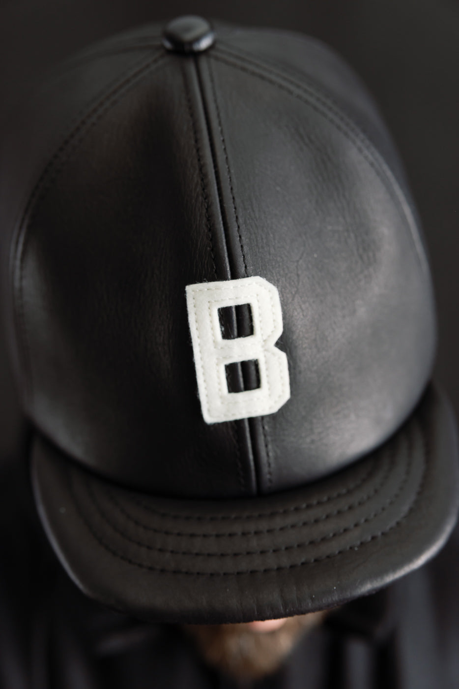 Close-up front view of black leather ball cap with white B patch, worn by male model, 5'9