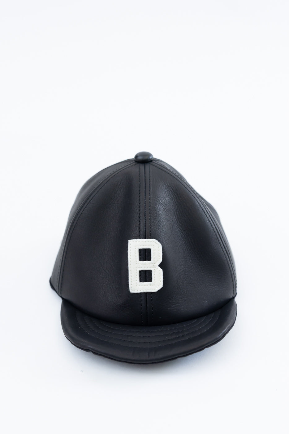 Front close-up of black leather baseball cap with white B patch, showing bill and panel seams