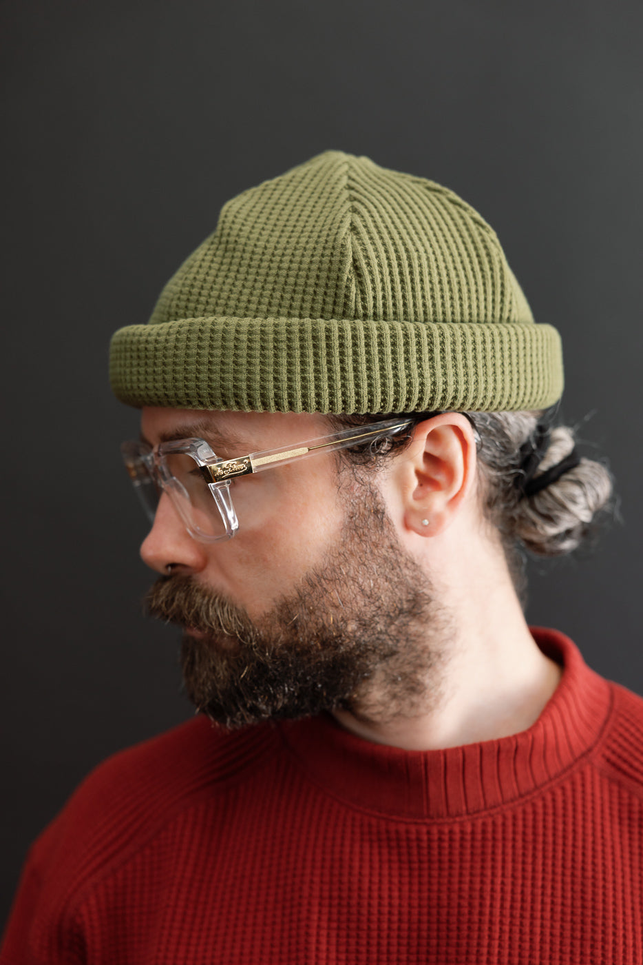Side profile close-up of a male model wearing an olive green waffle knit beanie and clear glasses.