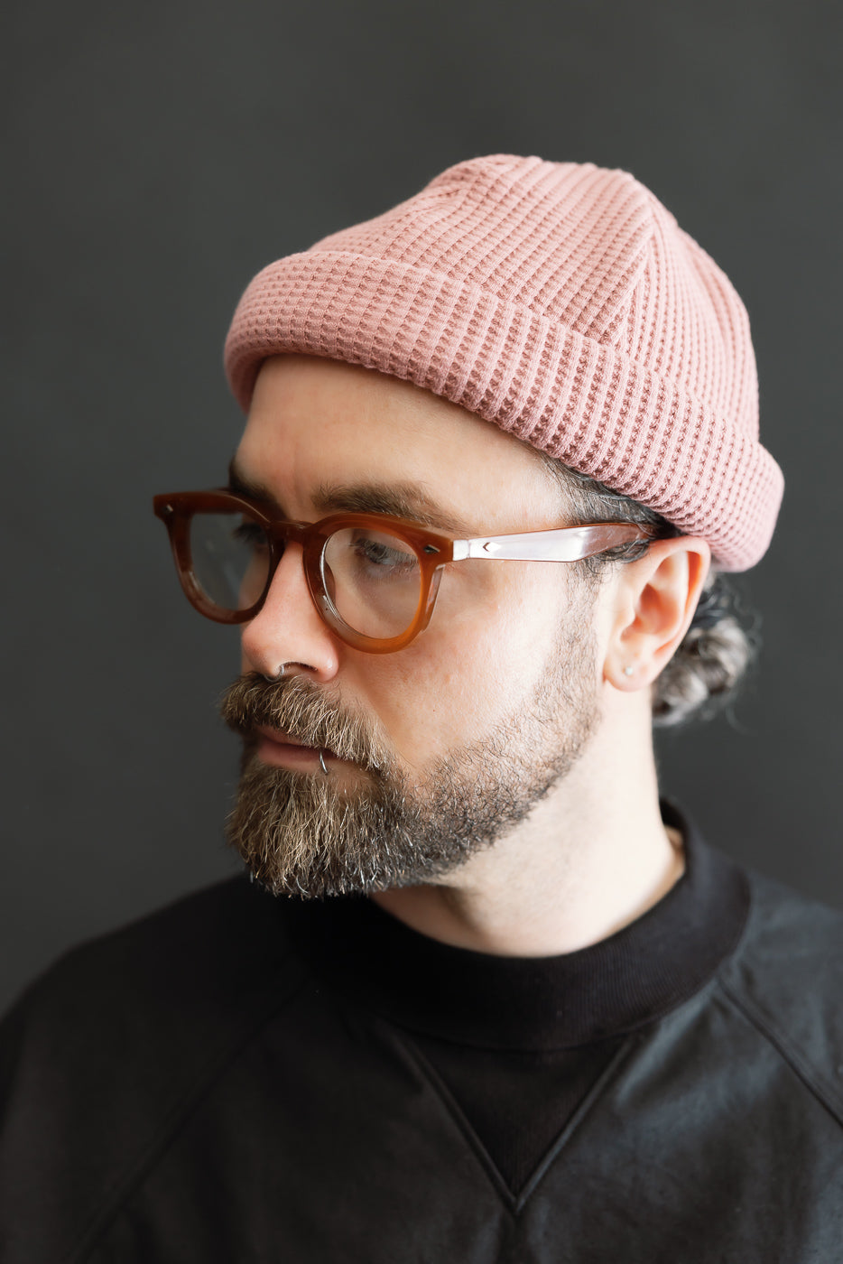 Man wearing JM6607 - Waffle Knit Cap - 448 Old Raspberry, a 100% cotton, pink waffle-knit beanie. The cap's textured knit and elongated design offer versatile styling, complementing his translucent eyeglasses and dark sweatshirt.
