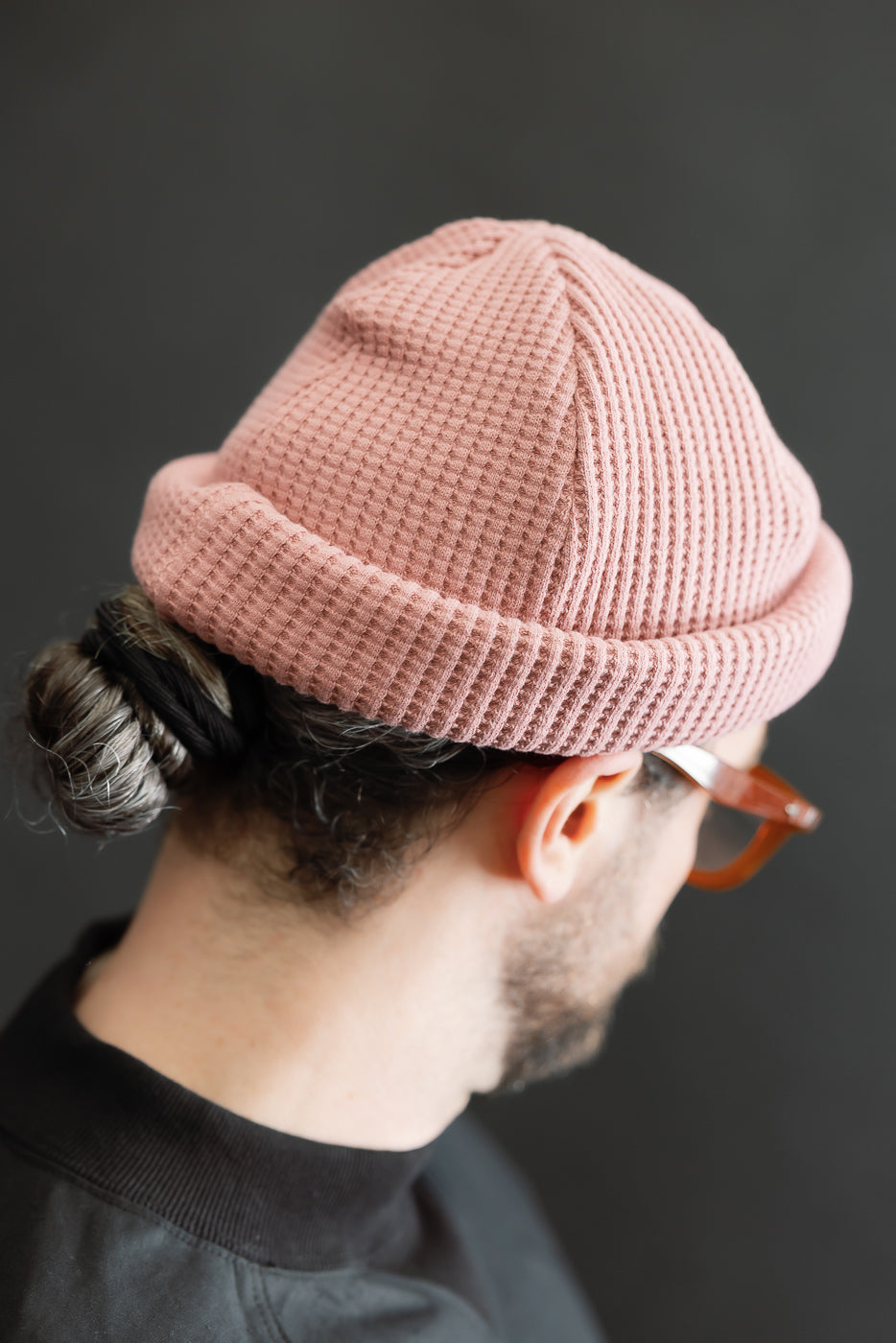 Back view of a person wearing the JM6607 - Waffle Knit Cap - 448 Old Raspberry, showcasing its pink waffle-knit texture and rolled hem, complemented by a twisted bun and brown-framed eyeglasses.