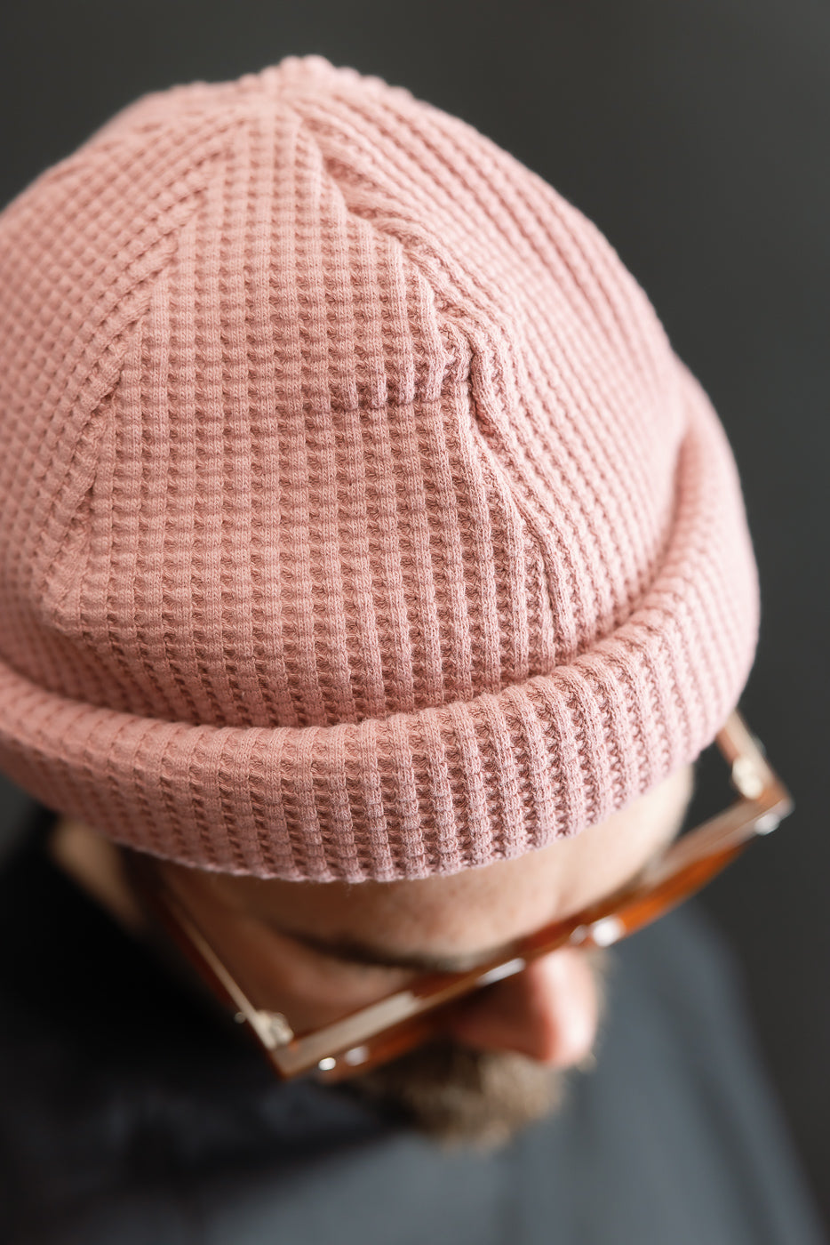 A person wearing the JM6607 - Waffle Knit Cap - 448 Old Raspberry, showcasing its rolled-up, waffle-knit texture and versatile fit, paired with tortoiseshell glasses, viewed from above against a dark background.