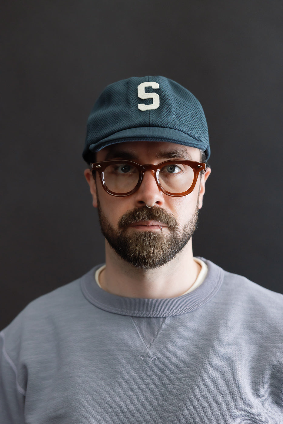Man wearing JM6623 - Sashiko BB Cap S - 280 Iron Navy, featuring textured blue-green sashiko fabric, round tortoiseshell glasses, and a light gray crewneck sweatshirt against a dark background.