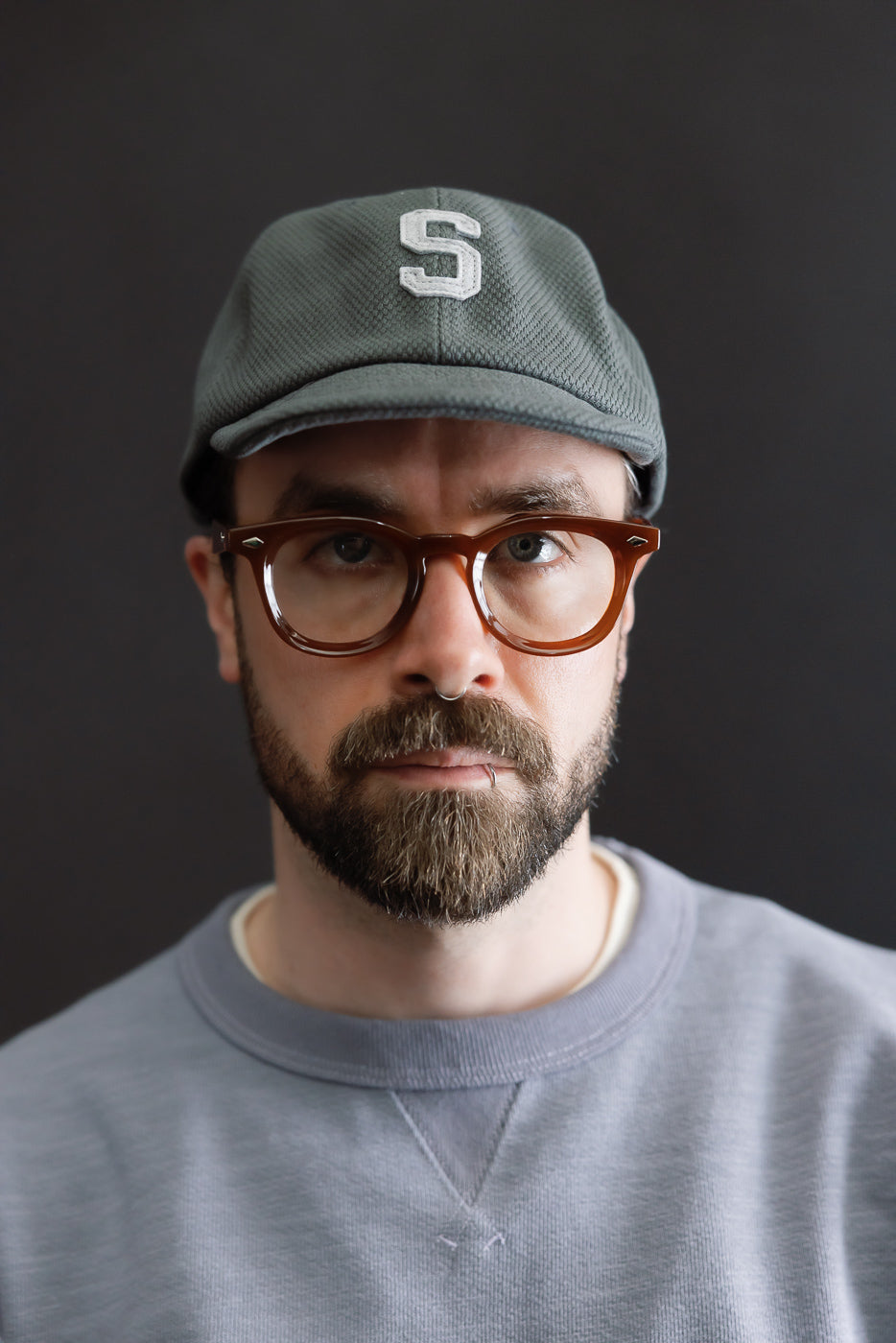 JM6623 - Sashiko BB Cap S features a textured sashiko fabric, highlighted by a raised white S on the front. The cap is modeled by a bearded man with glasses and facial piercings.