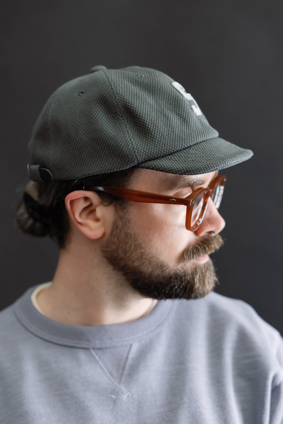 Side-profile of a man wearing the JM6623 - Sashiko BB Cap S - 333 Falcon Gray, showcasing its textured sashiko fabric and embroidered logo. Paired with amber-brown eyeglasses and a light grey sweatshirt.