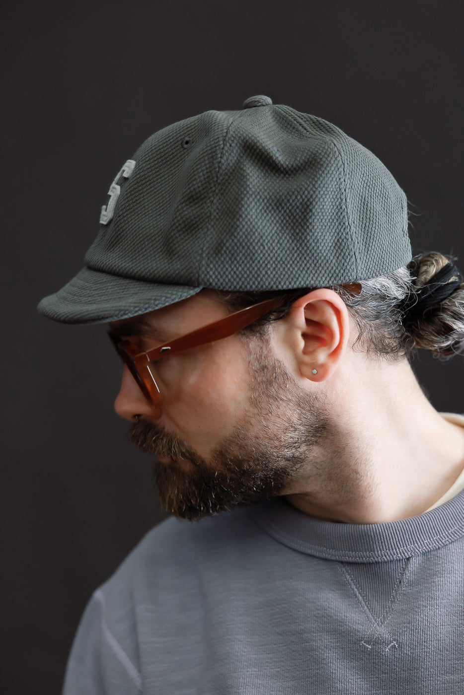 JM6623 - Sashiko BB Cap S - 333 Falcon Gray on model, showcasing its sashiko-textured fabric and small letter emblem, complemented by brownish-tinted eyeglasses and a slate-gray crewneck sweatshirt.
