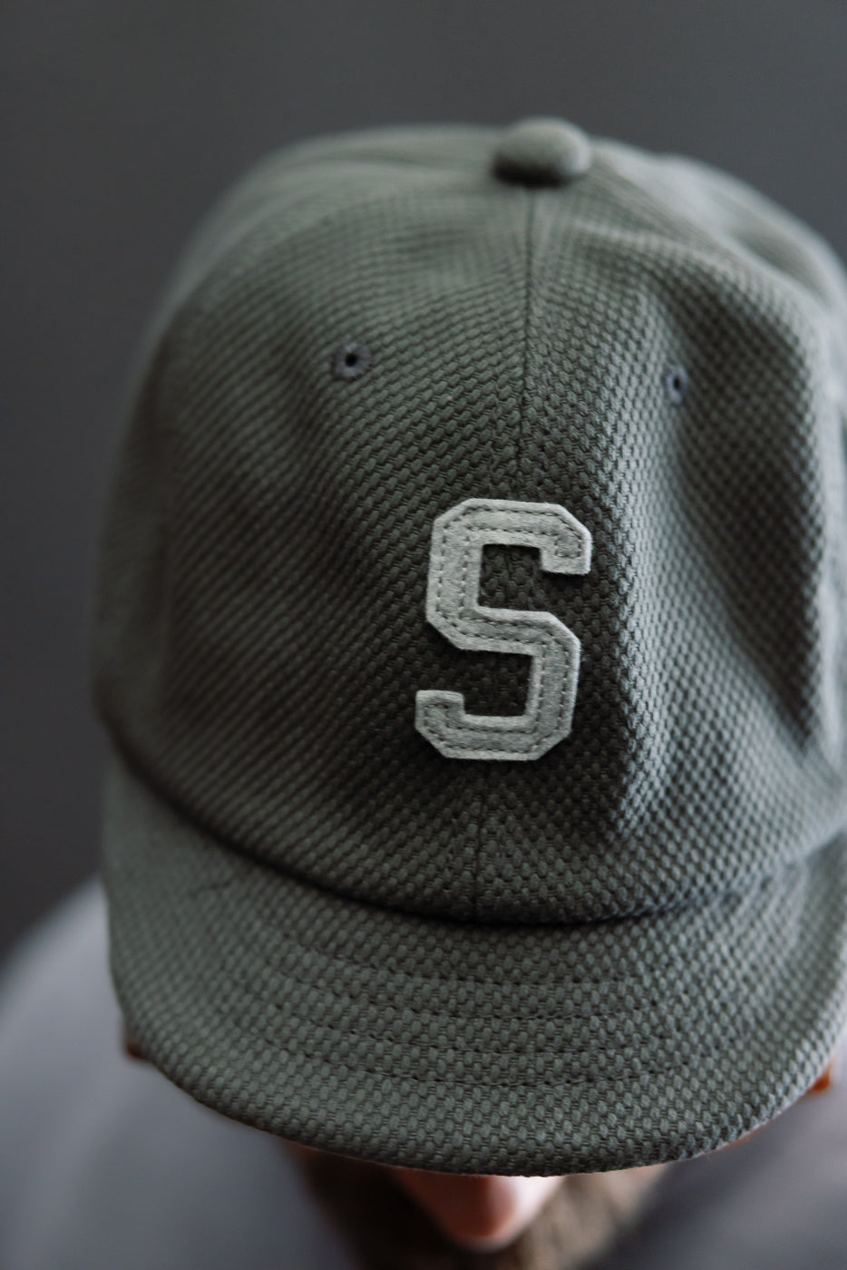 Close-up of JM6623 - Sashiko BB Cap S - 333 Falcon Gray, showcasing an olive-green textured cap with a raised light-grey S on the front, highlighting its unique sashiko fabric.