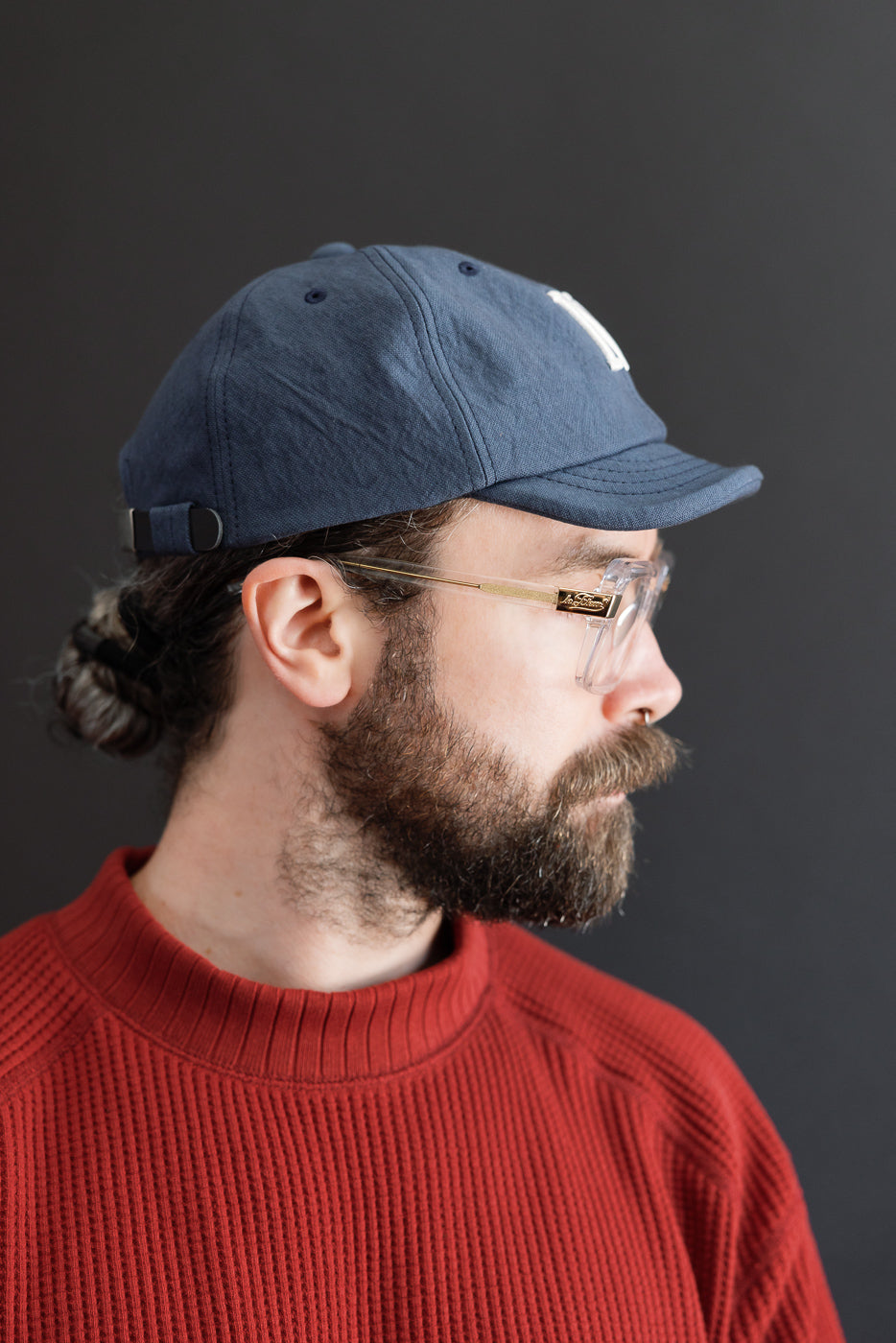 Side profile close-up of a male model wearing a navy blue baseball cap and red ribbed sweater.