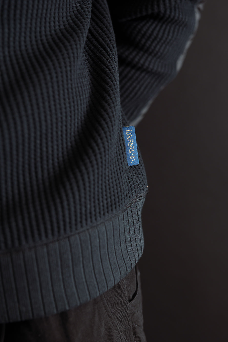 Close-up of a male model's side showing navy ribbed knit fabric, hem, and Lavenham tag.