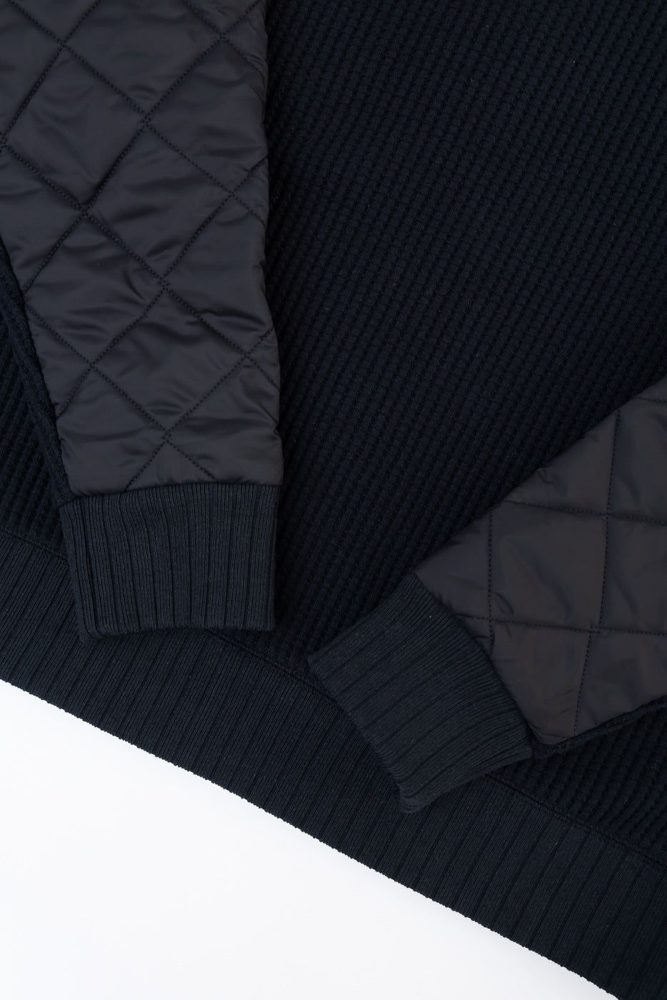 Close-up flat lay of the front of a black ribbed knit sweater with quilted nylon sleeves.