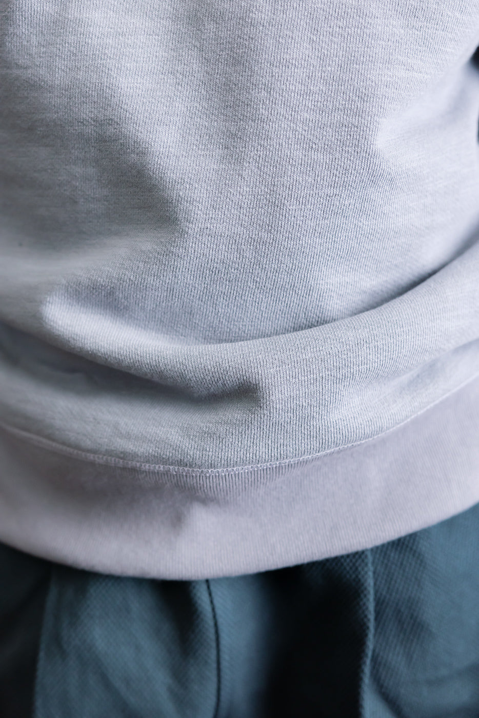 Close-up of the JM7292 - GG Sweat Crewneck's lower hem, showcasing its grainy texture and detailed knit, resting over greenish-teal pants, highlighting the sweatshirt's unique fabric weave and craftsmanship.