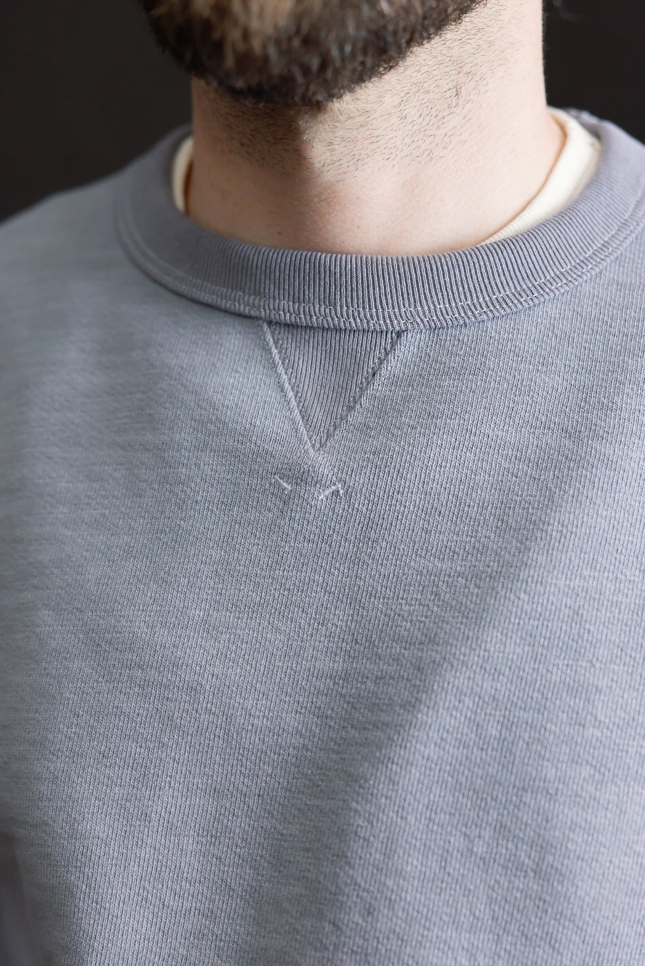 Close-up of a person wearing the JM7292 - GG Sweat Crewneck, highlighting the light gray texture and V-stitch detail below the collar, layered over a lighter T-shirt, illustrating the sweatshirt's weighty, grainy weave.