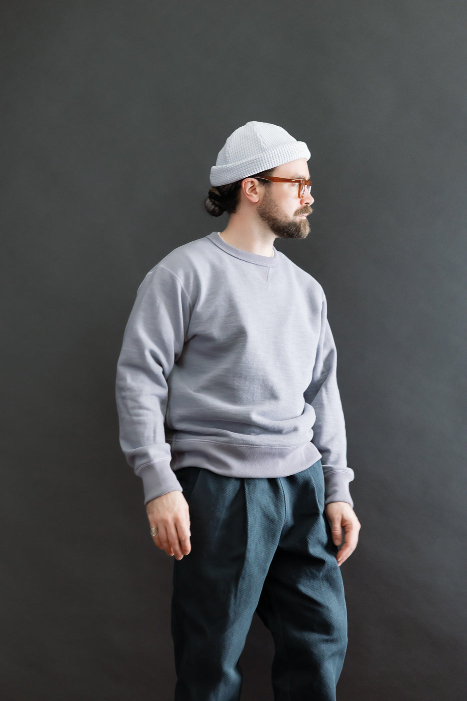 Male model wearing the JM7292 - GG Sweat Crewneck - 354 Dry Purple, showcasing its grainy texture and vintage-inspired design. Styled with pleated trousers, beanie, and glasses, exemplifying Dant's premium menswear aesthetic.