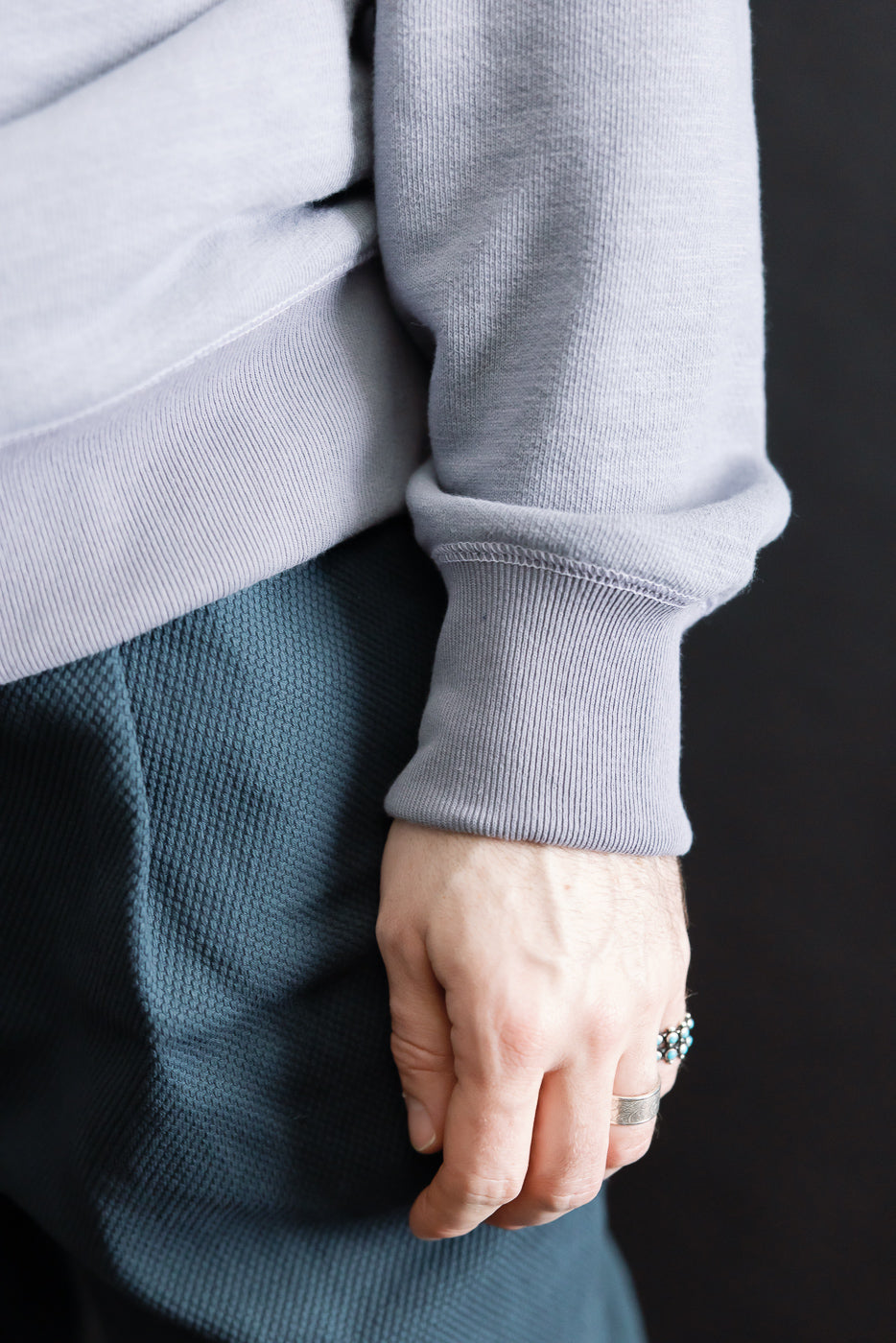 Close-up of a hand wearing the JM7292 - GG Sweat Crewneck, highlighting the light grey ribbed cuff and grainy texture, complemented by textured trousers and two rings.