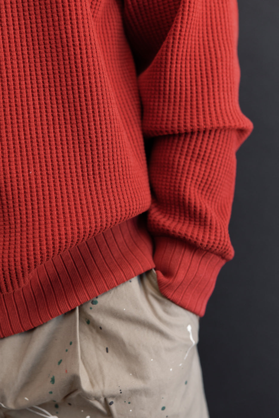 Close-up of the side and hem of a red waffle knit sweater and tan paint-splattered pants.