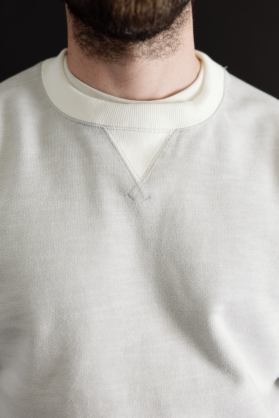 Close-up front view of a light grey crewneck sweatshirt showing ribbed collar and V-stitch detail.
