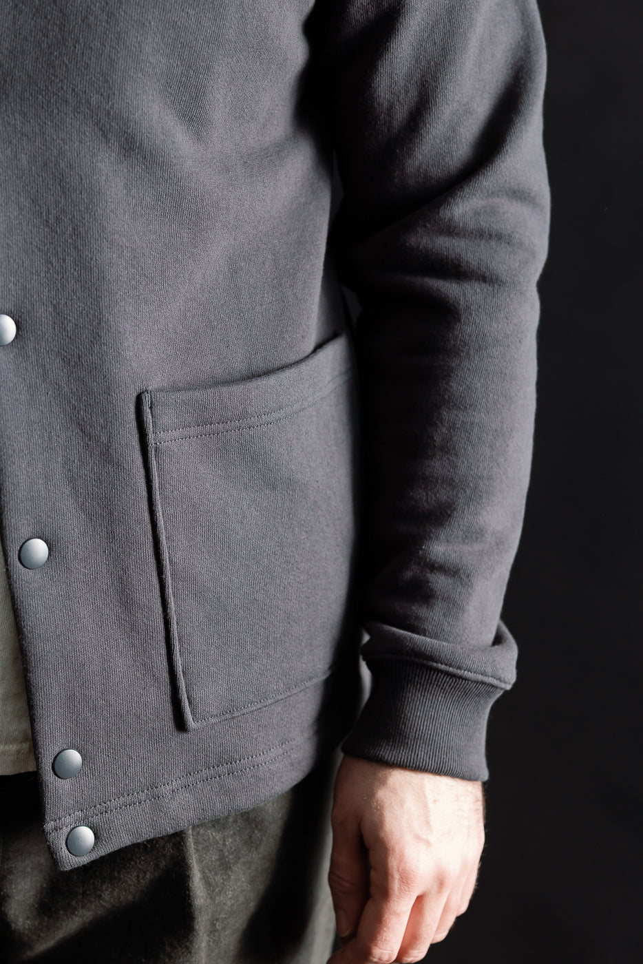 Close-up of front right side of men's black collarless jacket on male model, showing snap buttons, large pocket, and ribbed cuff. Model is 5'9