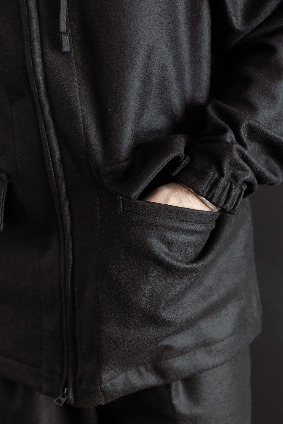 Close-up of male model's hand in front pocket of black wool jacket, showing pocket detail and fabric texture, front side view, model is 5'9