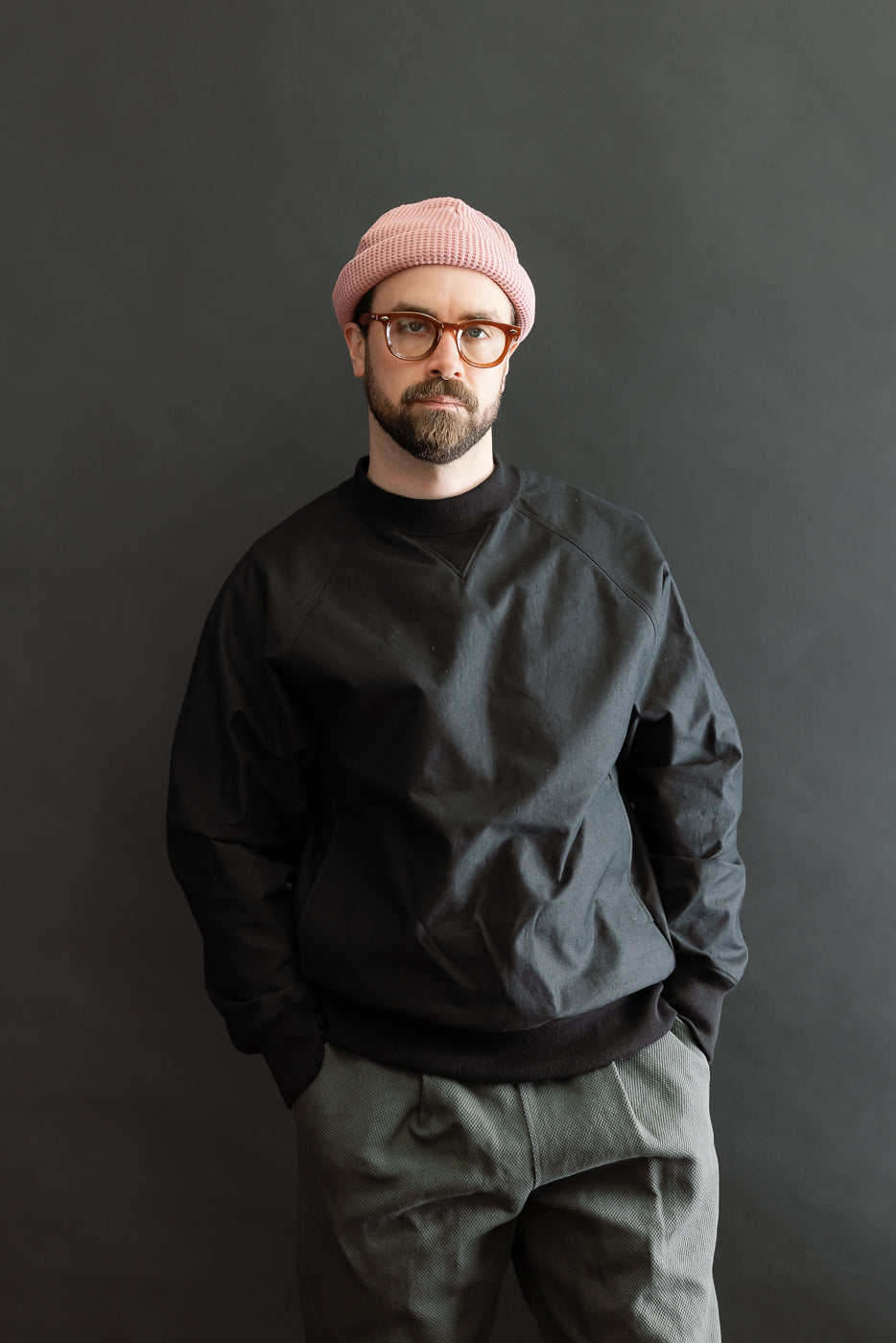 Model wearing JM8615 - BN Crewneck in black, showcasing its loose fit, raglan sleeves, and mid-neck with V-gusset. Hands in pockets, highlighting the pullover's casual style against a dark backdrop.