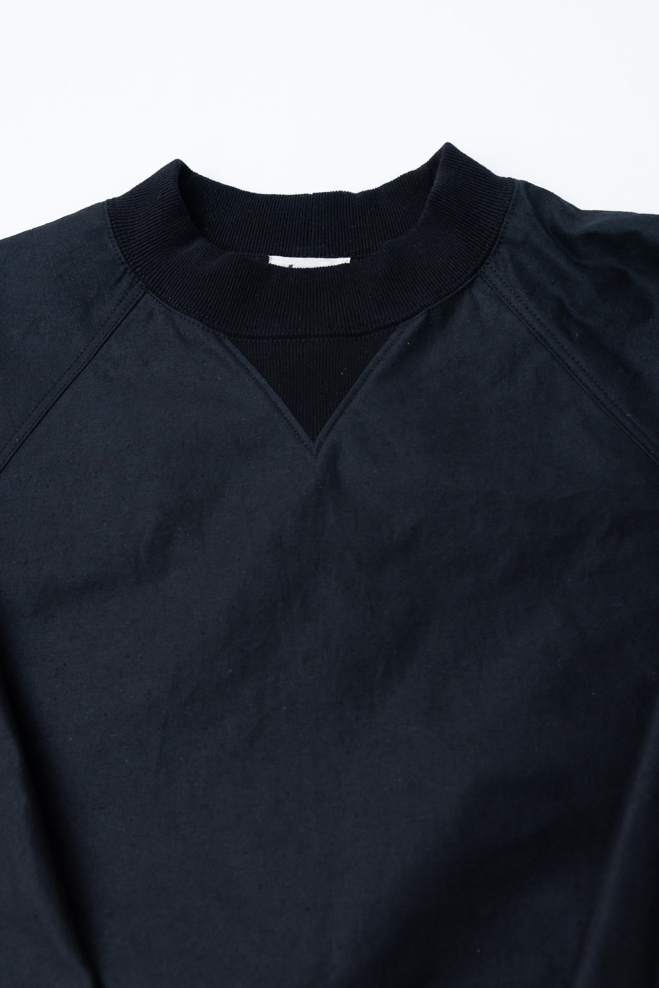 JM8615 - BN Crewneck - 07 Black: A dark crewneck sweatshirt featuring ribbed collar, raglan shoulder seams, and a V-shaped stitch detail below the neckline, emphasizing a rustic, textured finish.