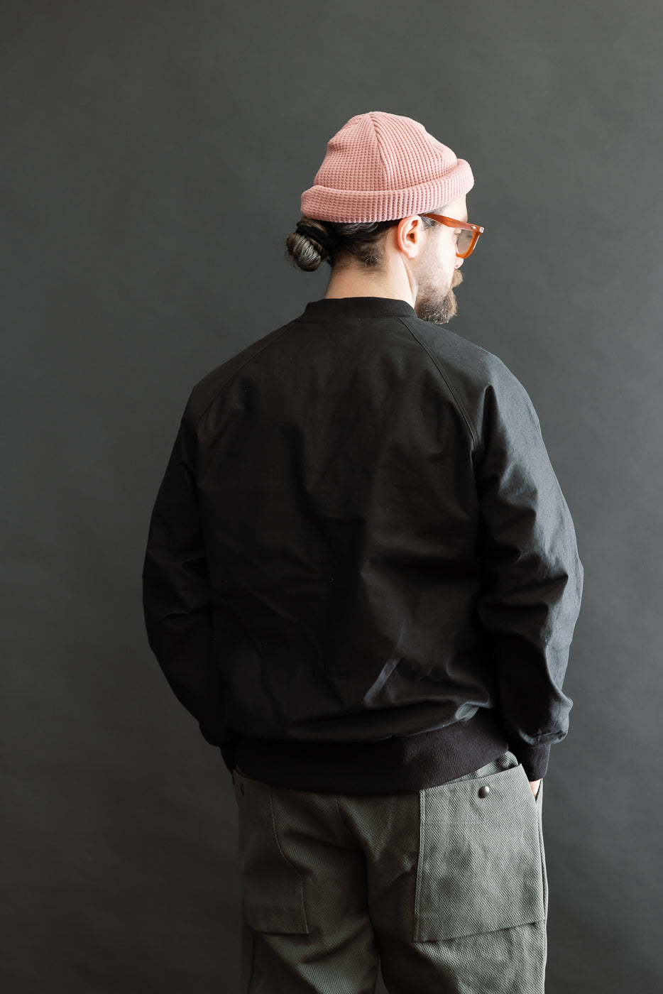 Person wearing the JM8615 - BN Crewneck in black, featuring a loose fit with raglan sleeves and a mid-neck with V-gusset, styled with olive cargo pants and a pink beanie.
