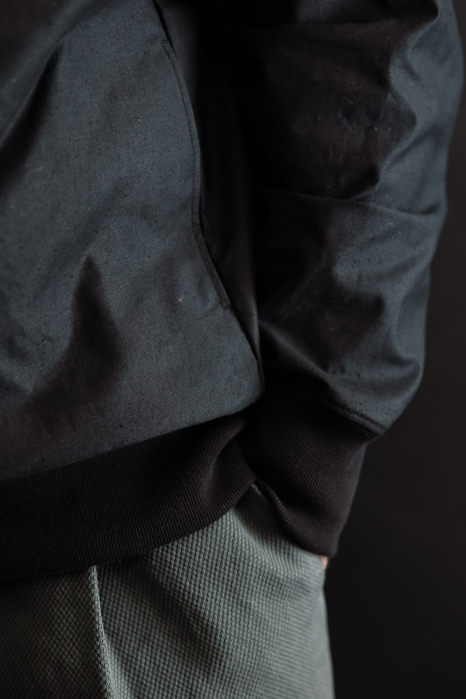 Close-up of a person wearing the JM8615 - BN Crewneck - 07 Black, focusing on the ribbed hem of the dark jacket and seam pockets, paired with muted-green textured trousers.