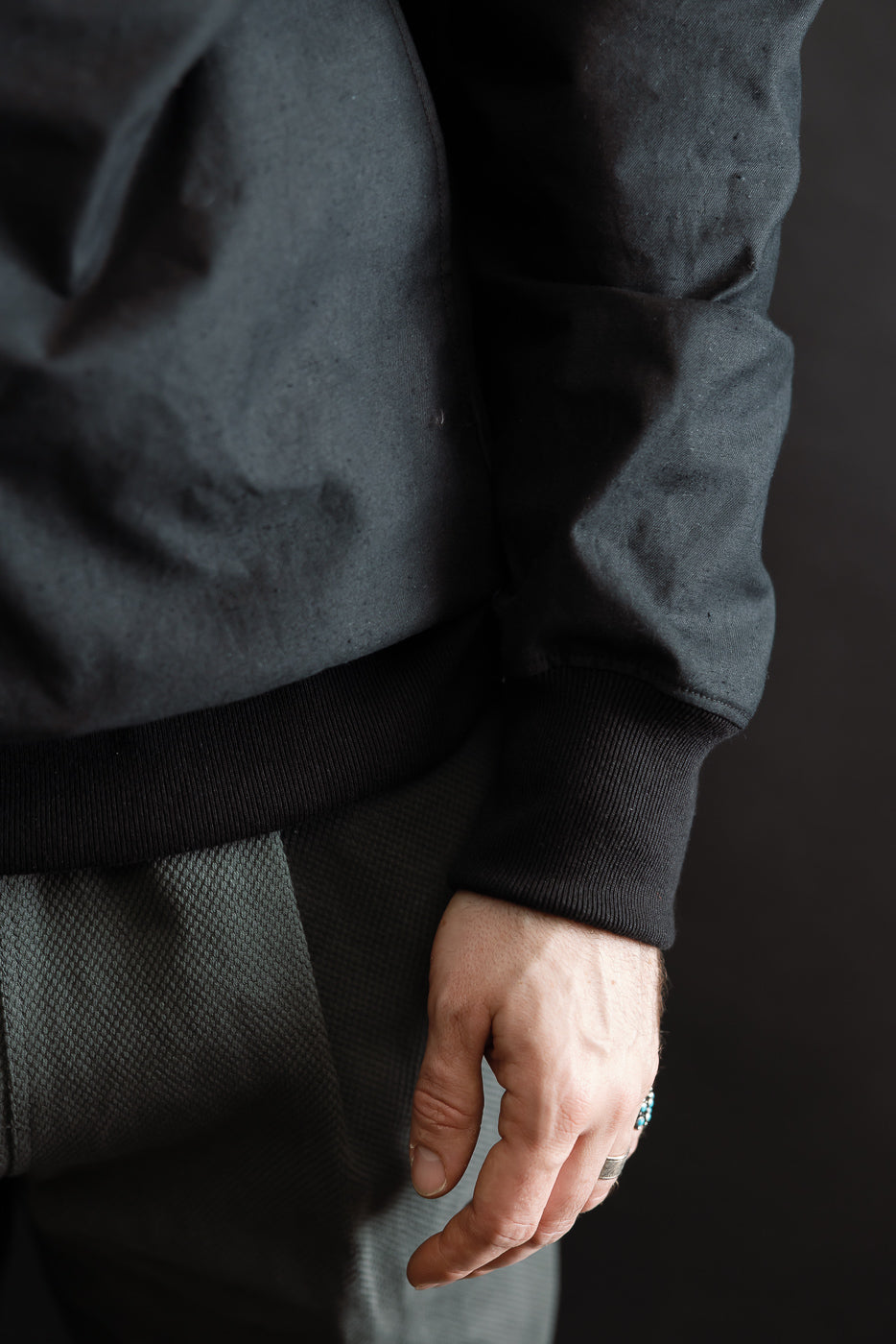 JM8615 - BN Crewneck - 07 Black shown worn, highlighting the ribbed hem and cuff of the black sweatshirt and textured dark-gray pants, with a relaxed hand adorned by two rings.