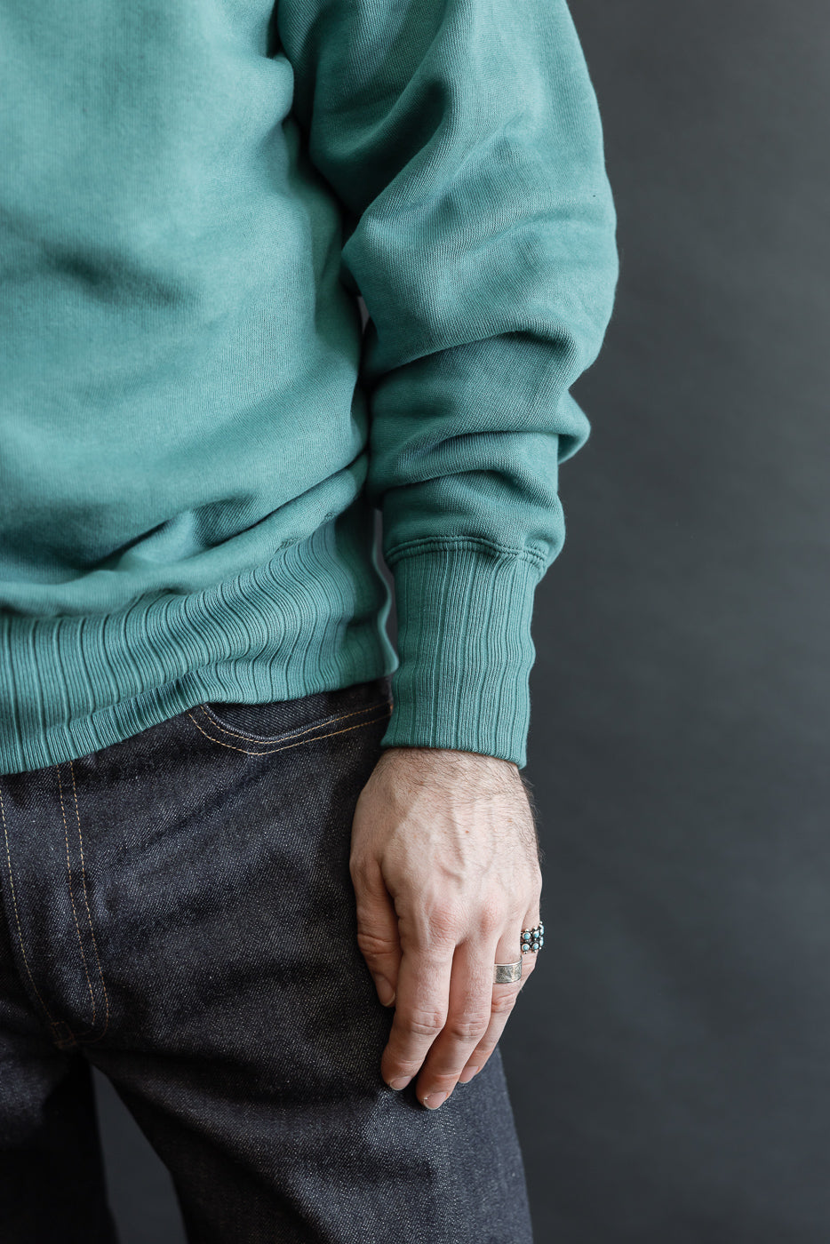 JG-CS24 - The Albert Sweat - Blue Green: A teal-green sweatshirt with ribbed hem and cuffs, worn with dark denim jeans, showcasing a vintage-inspired, seamless design and freedom sleeve for classic comfort.