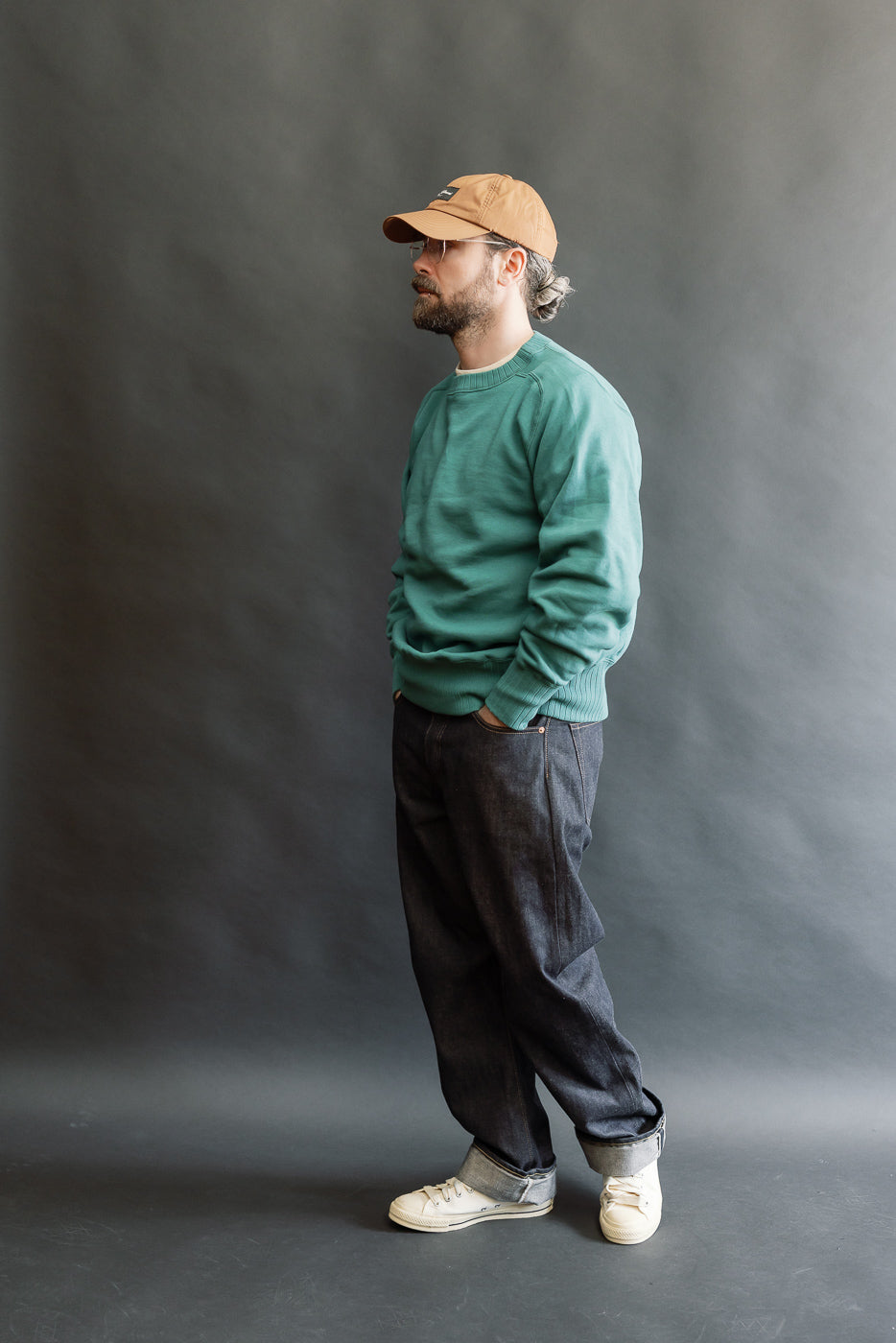 A man models the JG-CS24 Albert Sweat in blue green, showcasing its relaxed, seam-free silhouette with freedom sleeves, paired with dark denim jeans and canvas sneakers.