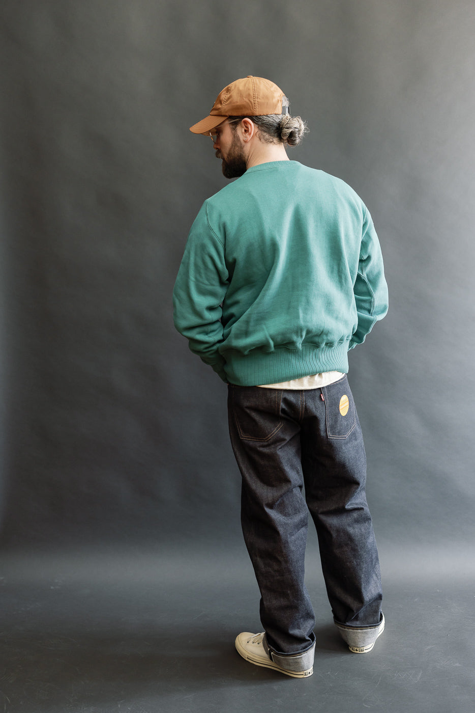 An adult male models the JG-CS24 Albert Sweat in blue-green, showcasing its relaxed fit and heritage-inspired design with freedom sleeve construction, paired with dark jeans and canvas sneakers.