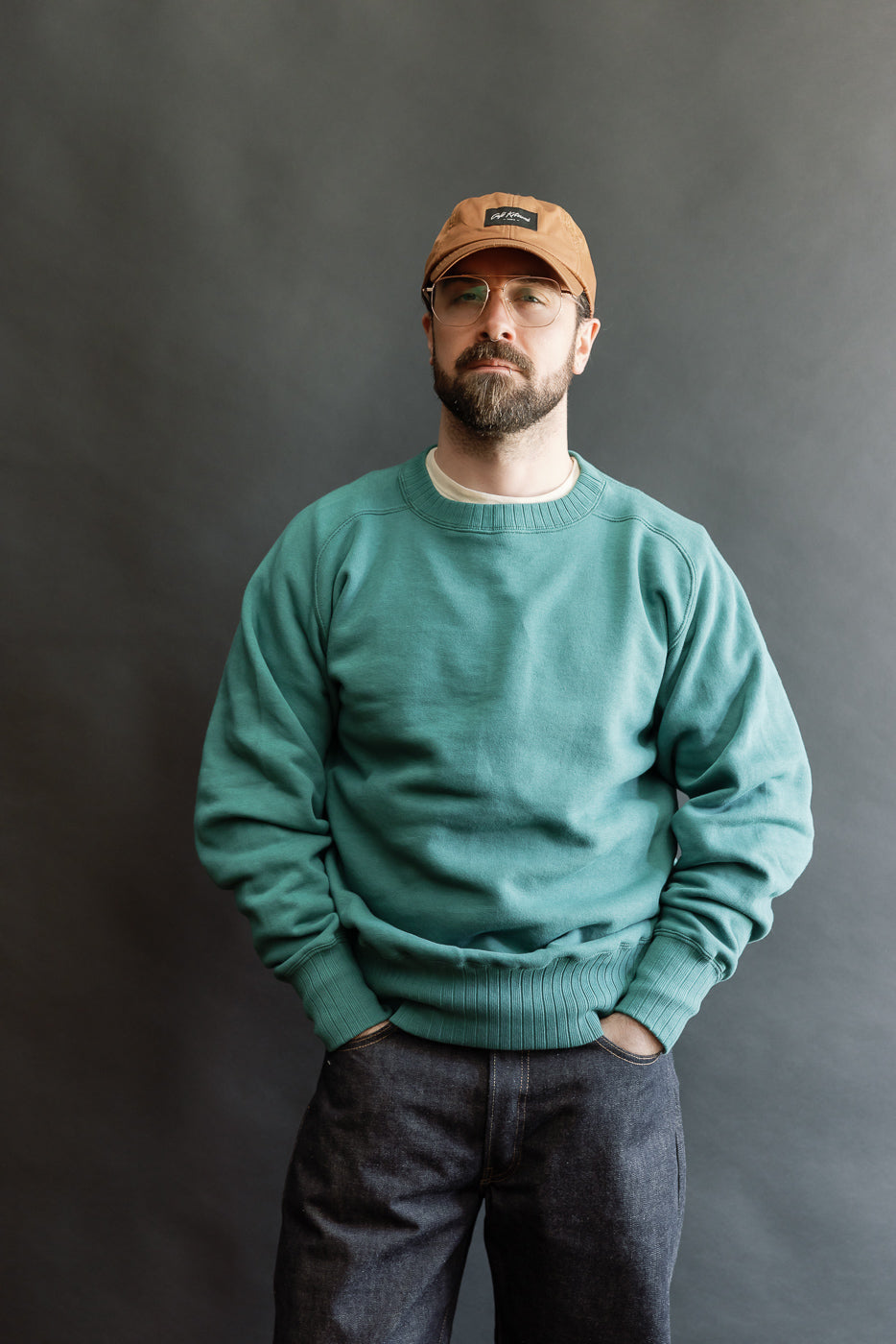 Man wearing JG-CS24 - The Albert Sweat - Blue Green stands confidently, showcasing its relaxed fit and heritage style. The teal crewneck complements dark denim, embodying Dant's high-end, heritage-inspired aesthetic.