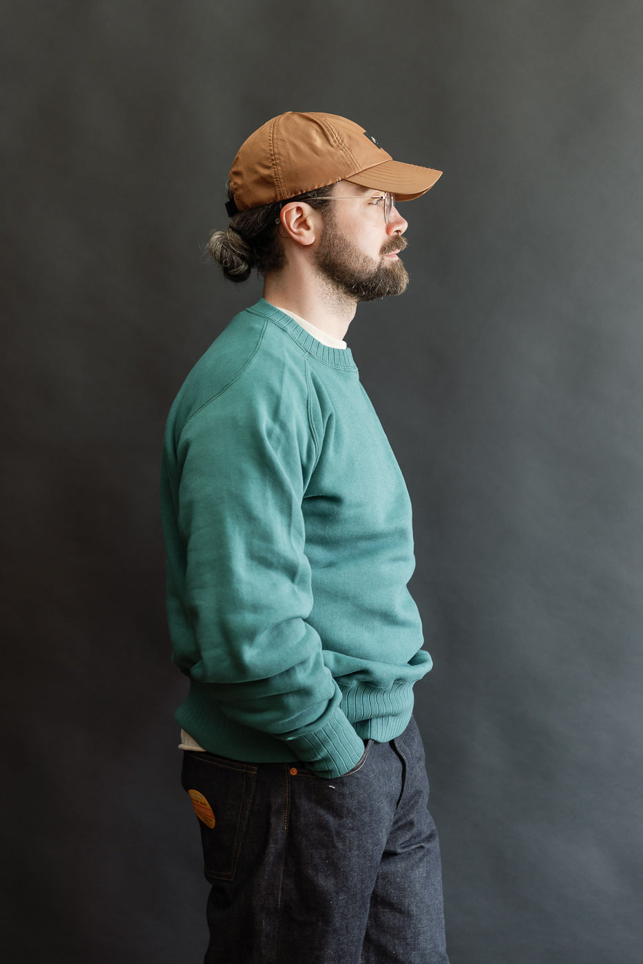 Man wearing JG-CS24 - The Albert Sweat - Blue Green, showcasing its relaxed fit and freedom sleeve design, inspired by Albert Einstein's 1940s sportswear, with hands in pockets, against a dark background.