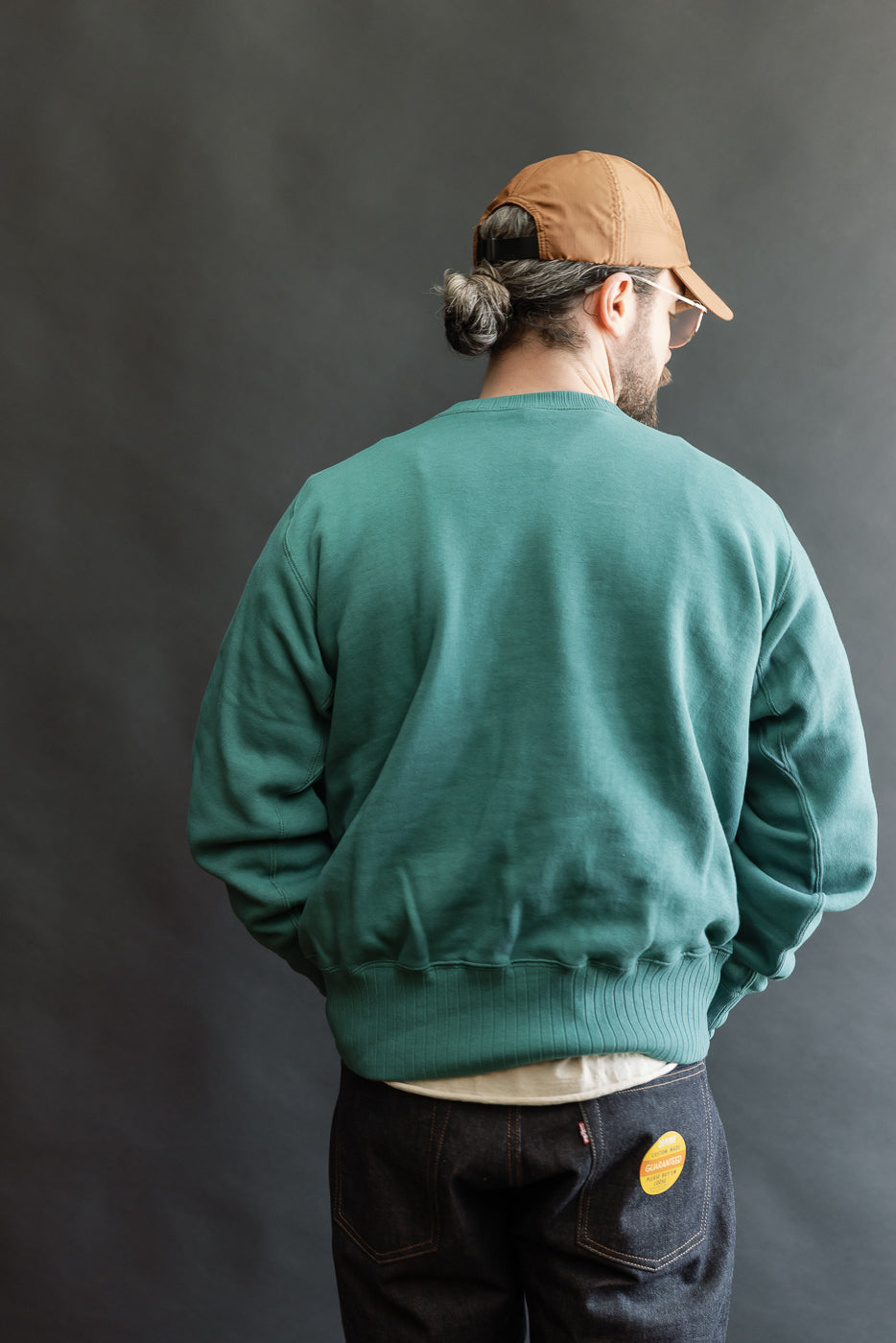 Model wearing the JG-CS24 Albert Sweat in teal-green, showcasing its seamless, relaxed drape and freedom sleeve construction, paired with dark blue jeans featuring a yellow patch on the back pocket.