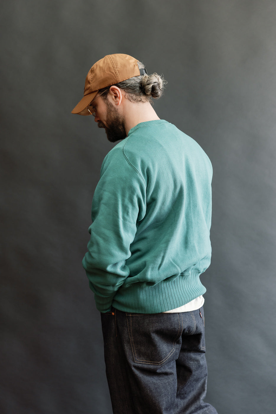 Bearded man in teal JG-CS24 - The Albert Sweat, inspired by Einstein’s 1940s sweatshirt, featuring circular knit, freedom sleeves, and no side seams, styled with low baseball cap and jeans.