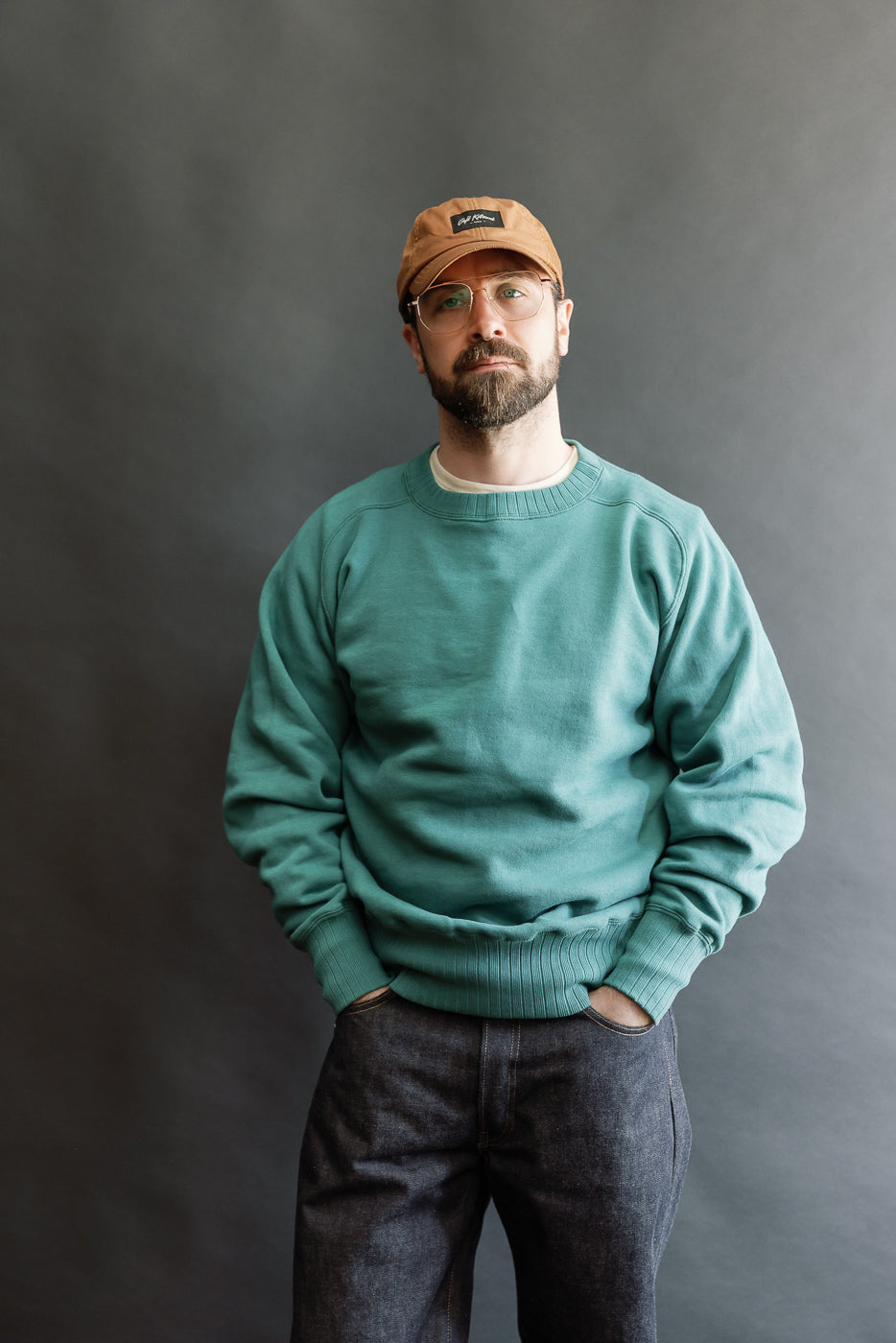 The JG-CS24 - The Albert Sweat - Blue Green, styled on a 5'9 Caucasian male, features a teal crewneck design with a relaxed fit, worn over a white T-shirt and dark jeans.